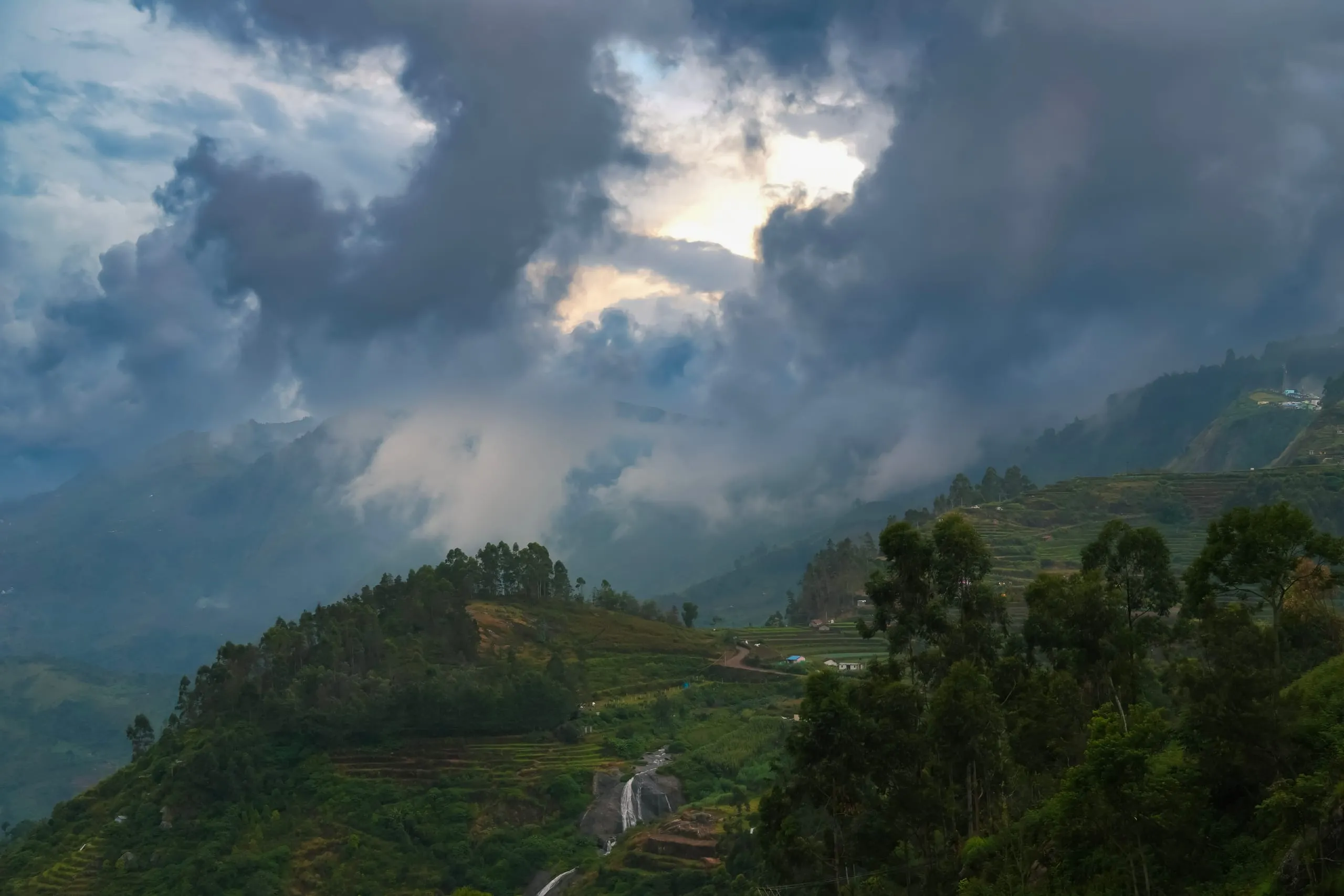 Heavy grey cloud breaking over a terraced hillside, with a waterfall and a stand of eucalyptus below