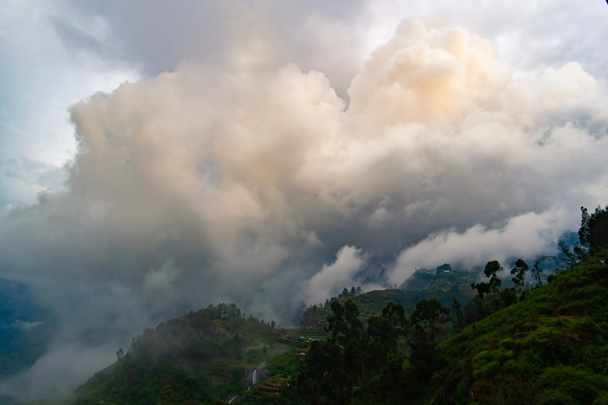 A large cumulus cloud spilling over a ridge and down into the valley, terraced fields and a waterfall beneath it