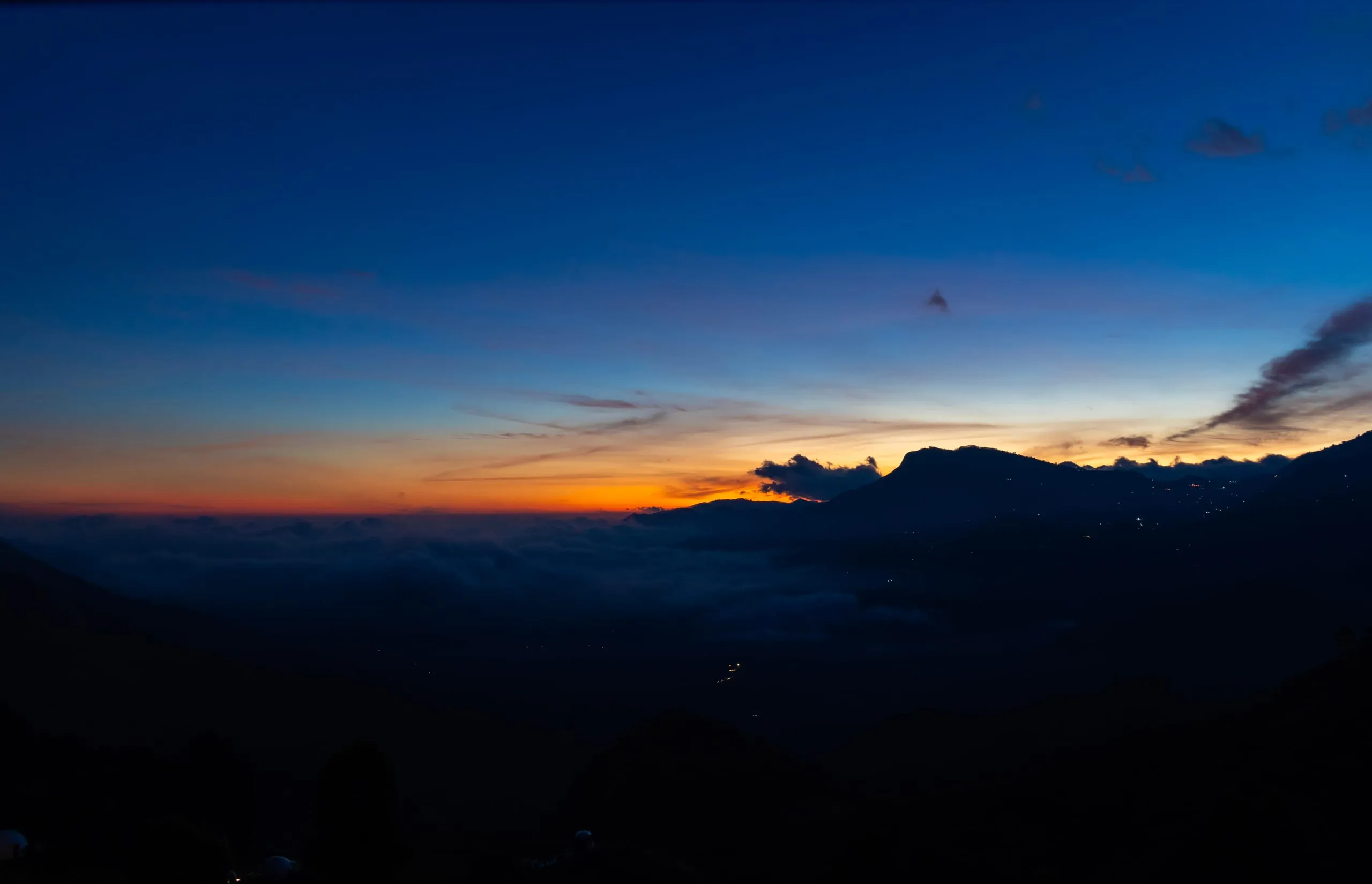 A band of orange on the horizon at dusk, cloud lying in the valley below and village lights along a dark ridge